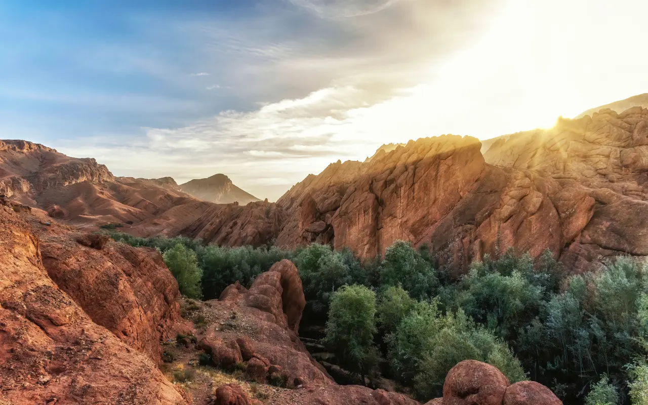 Paisaje del Alto Atlas al atardecer con formaciones rocosas rojizas y un oasis de vegetación frondosa entre las montañas.