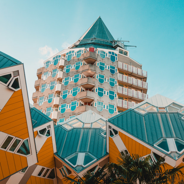 Vista de los icónicos edificios cúbicos amarillos y azules de Rotterdam en primer plano, con un edificio residencial de forma octogonal y techo puntiagudo al fondo, bajo un cielo claro y azul.