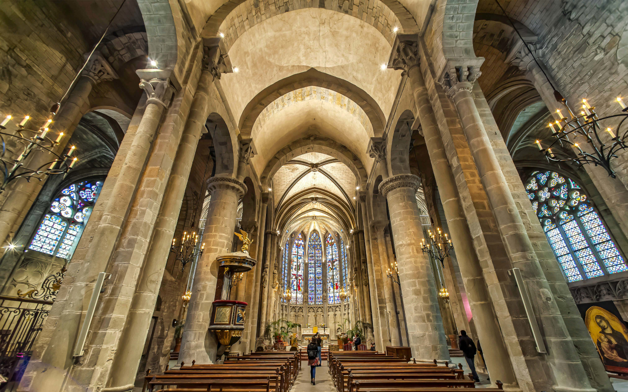 Interior gótico de la Basílica de San Nazario en Carcasona, con vitrales, columnas y bancos de madera.