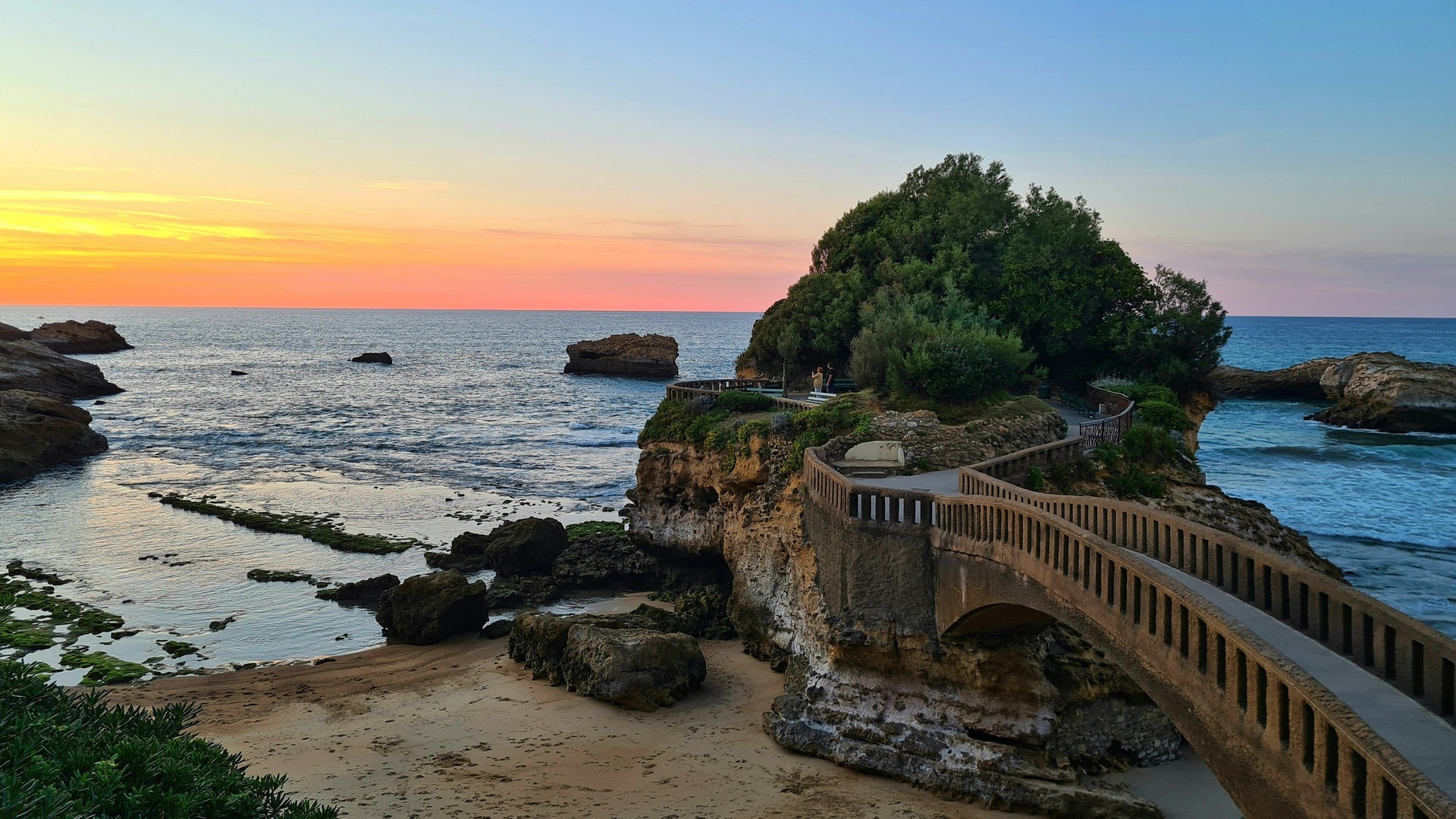 Atardecer en Biarritz, Francia, con el puente de piedra que conecta la costa con la Rocher de la Vierge, mar azul.