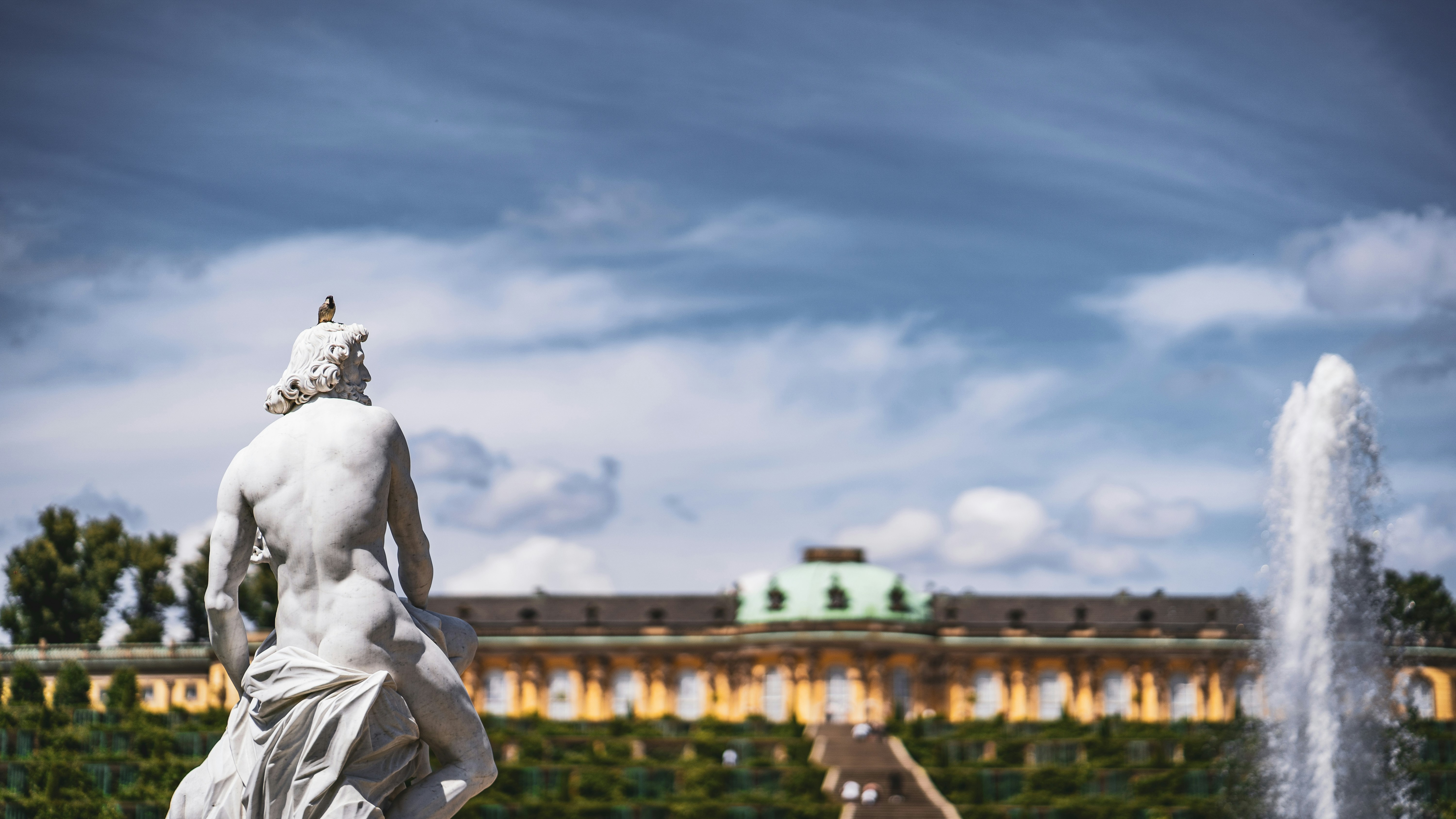 Estatua clásica con un pájaro en el Jardín de Sanssouci, Potsdam. Al fondo se ve la fuente y el Palacio de Sanssouci.