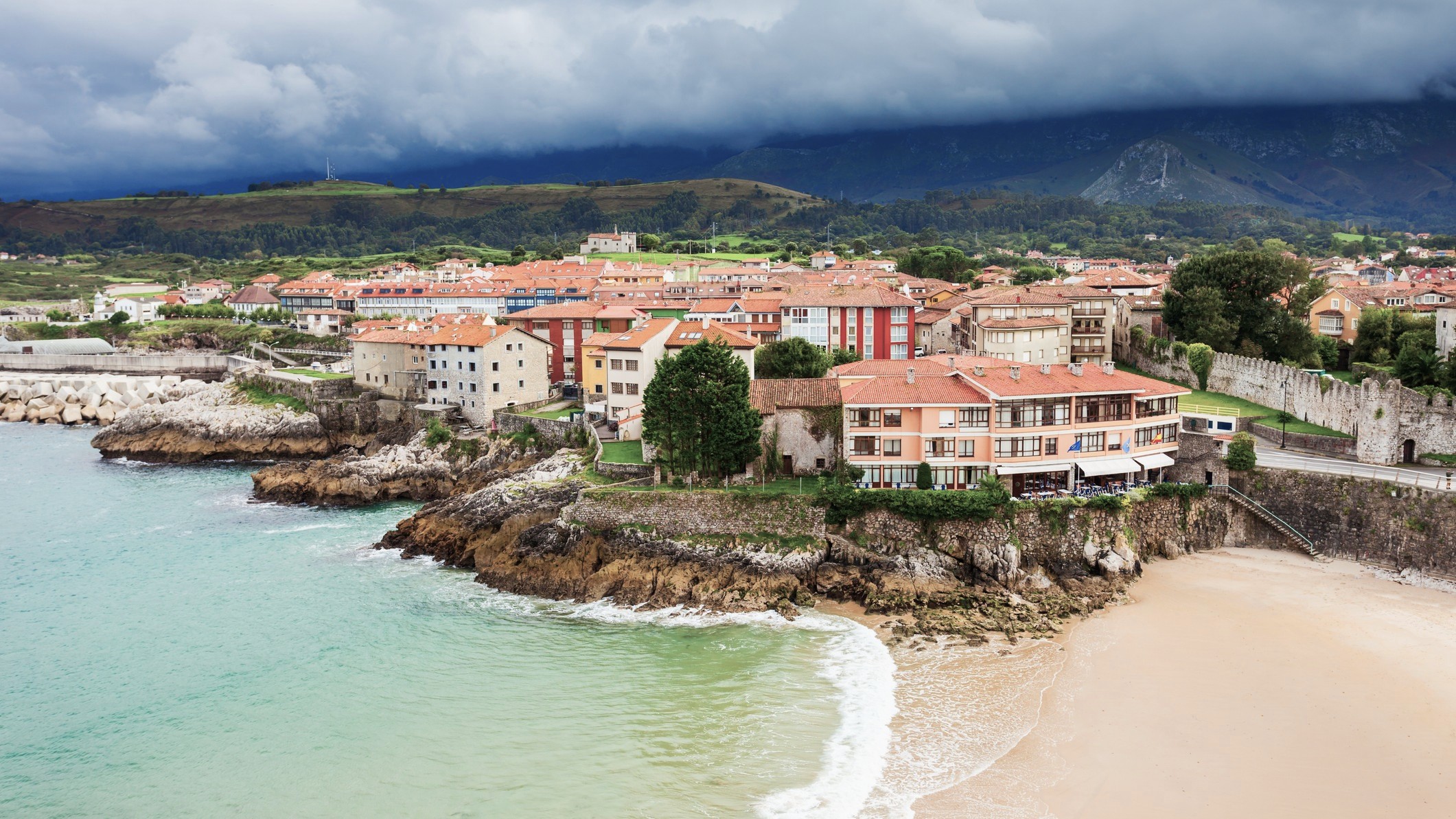 Vista de la villa costera de Llanes en Asturias, con casas sobre acantilados, playa dorada y montañas bajo nubes oscuras.
