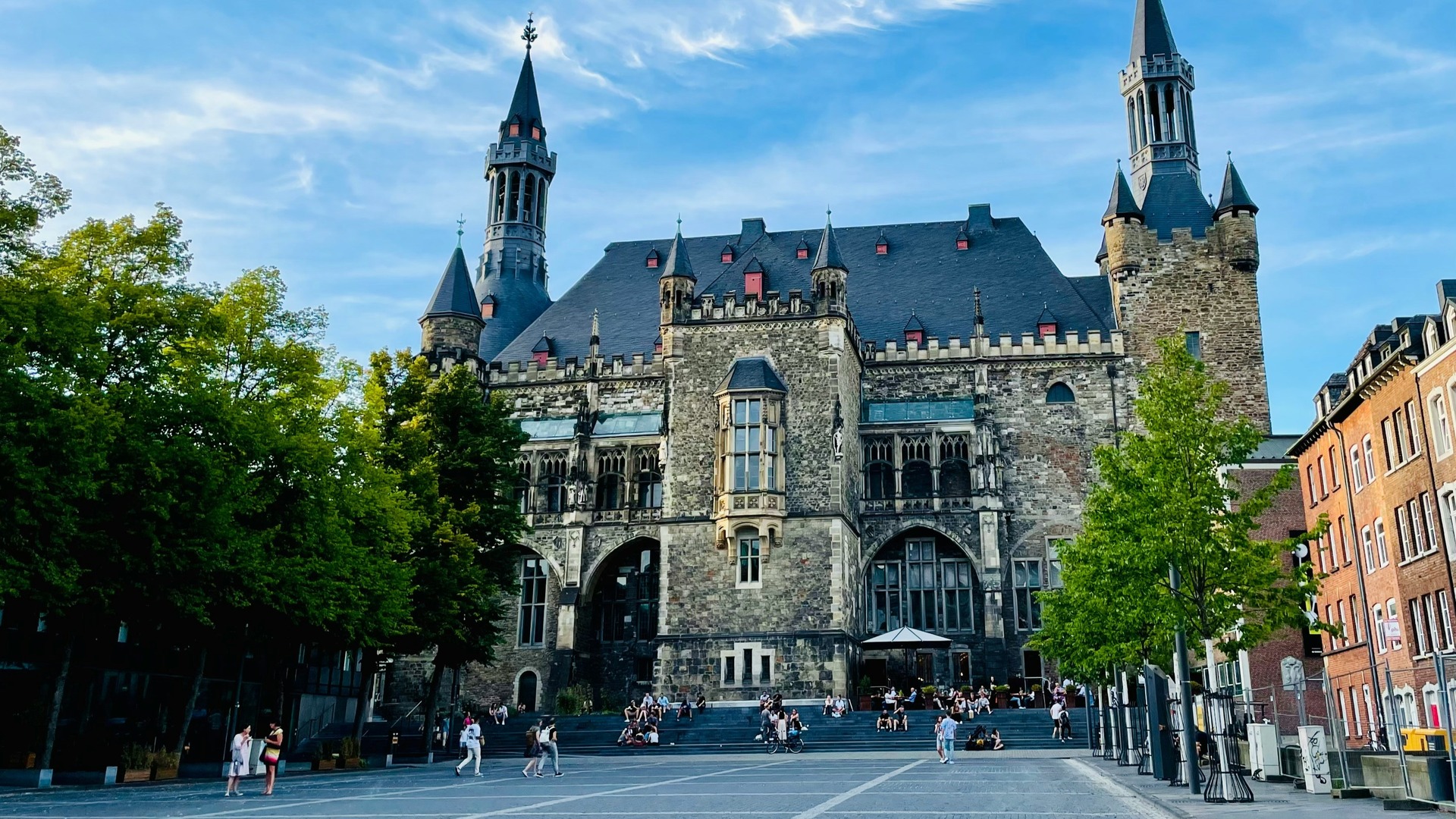 Edificio gótico y neoclásico del Ayuntamiento de Aquisgrán (Aachener Rathaus) con torres, en una plaza soleada.