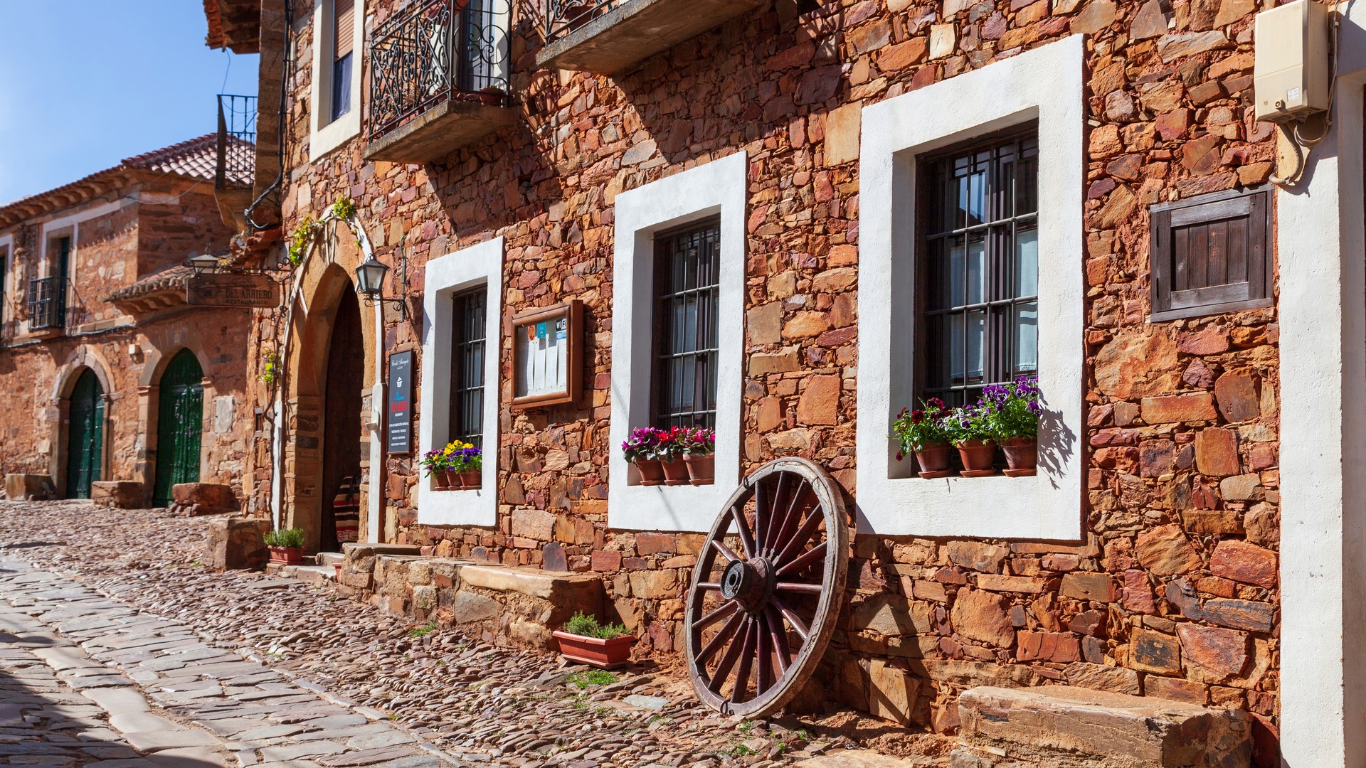 Calle de Castrillo de los Polvazares, León, con casa maragata de piedra rojiza, ventanas encaladas y rueda de carro.
