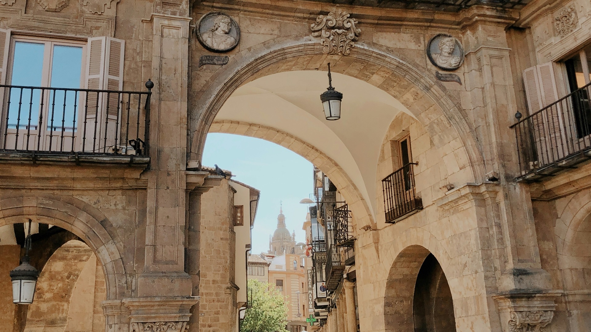 Arco de piedra en el centro histórico de Salamanca, con bustos y faroles colgantes, y la cúpula de la Catedral al fondo.