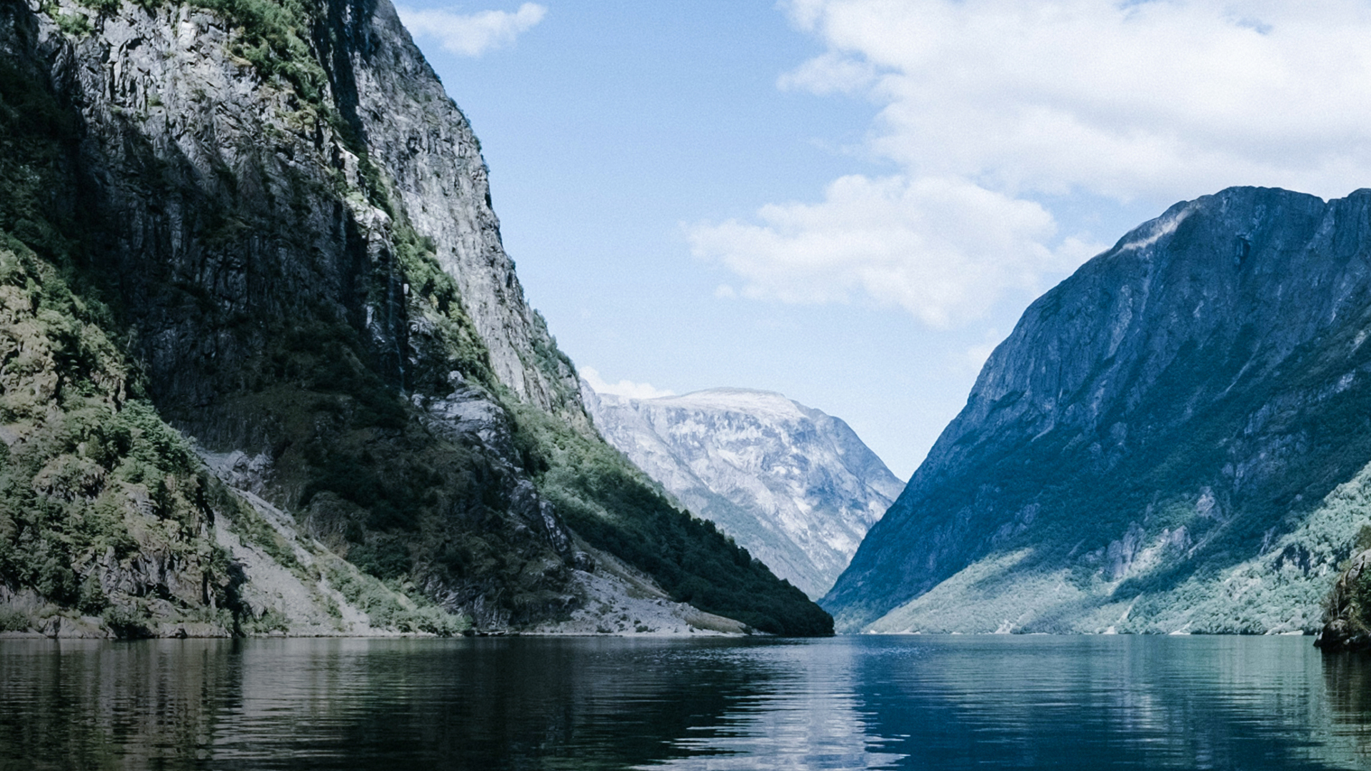Fiordo noruego rodeado de altas montañas y vegetación, con aguas tranquilas reflejando el cielo azul.
