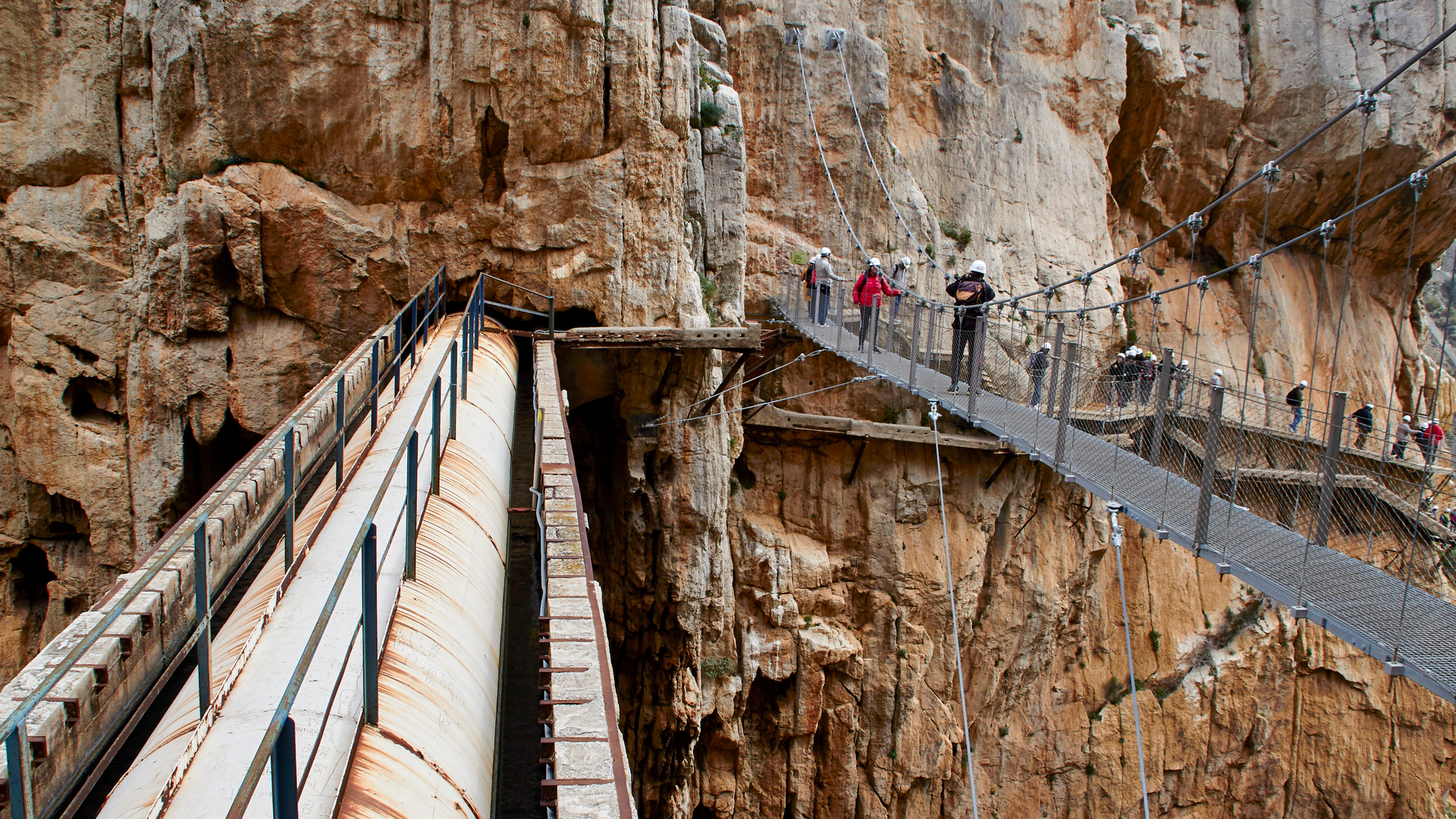 Senderistas cruzan puente colgante y tubería en el Caminito del Rey, Málaga.