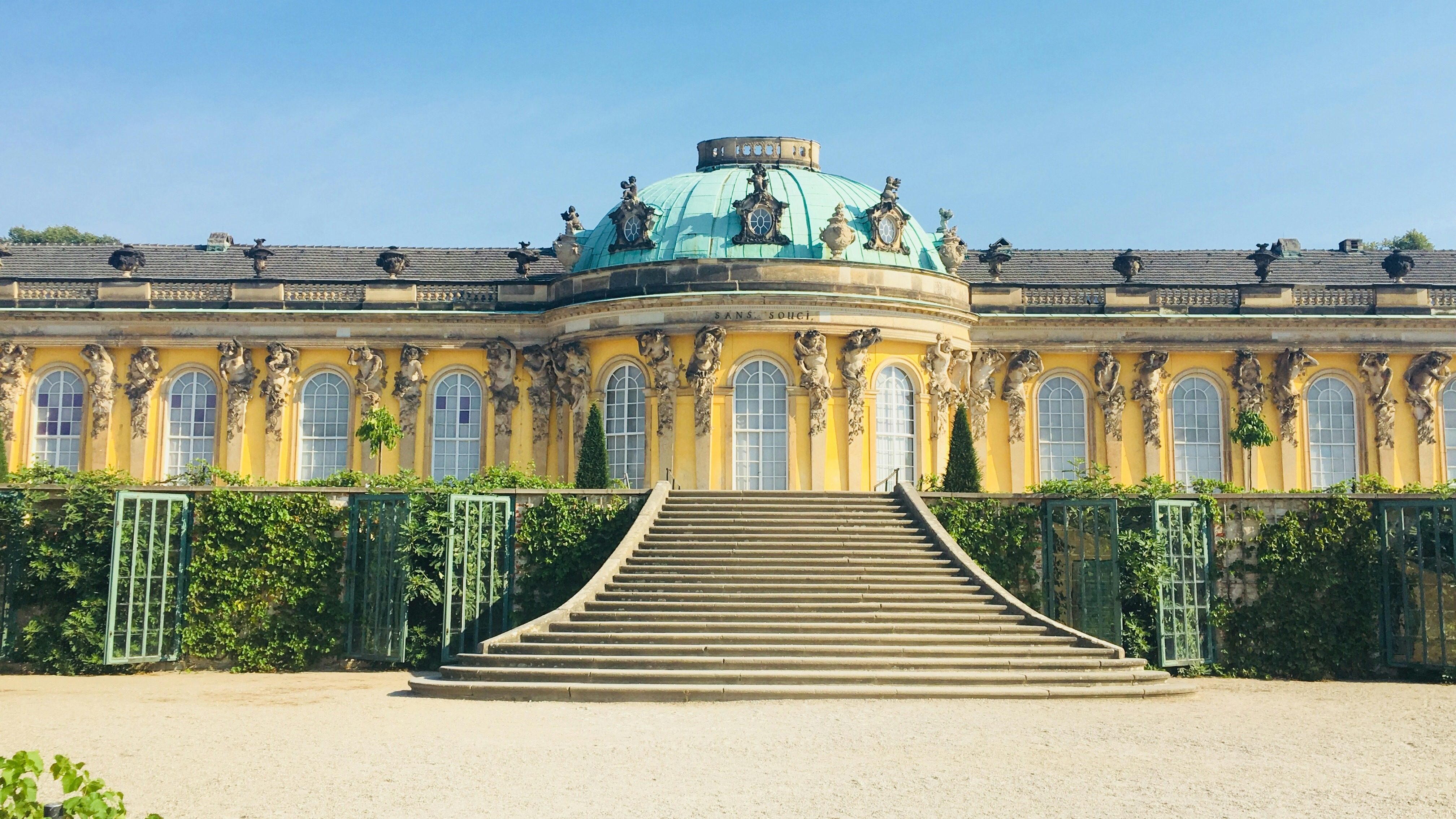 Gran escalinata que sube al Palacio de Sanssouci en Potsdam, Alemania, destacando su fachada amarilla y cúpula verde.