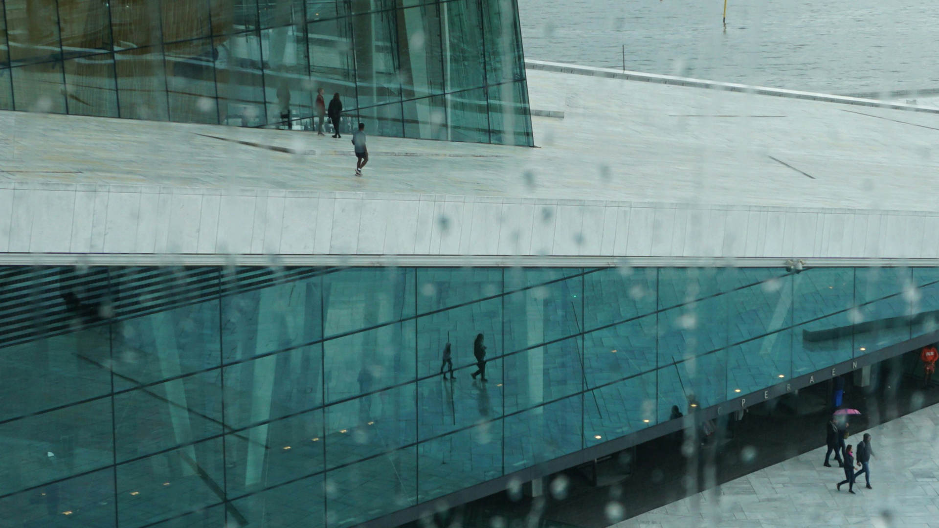 Reflejo en los cristales de la Ópera de Oslo, Noruega, con techo de mármol blanco y personas paseando bajo la lluvia. (