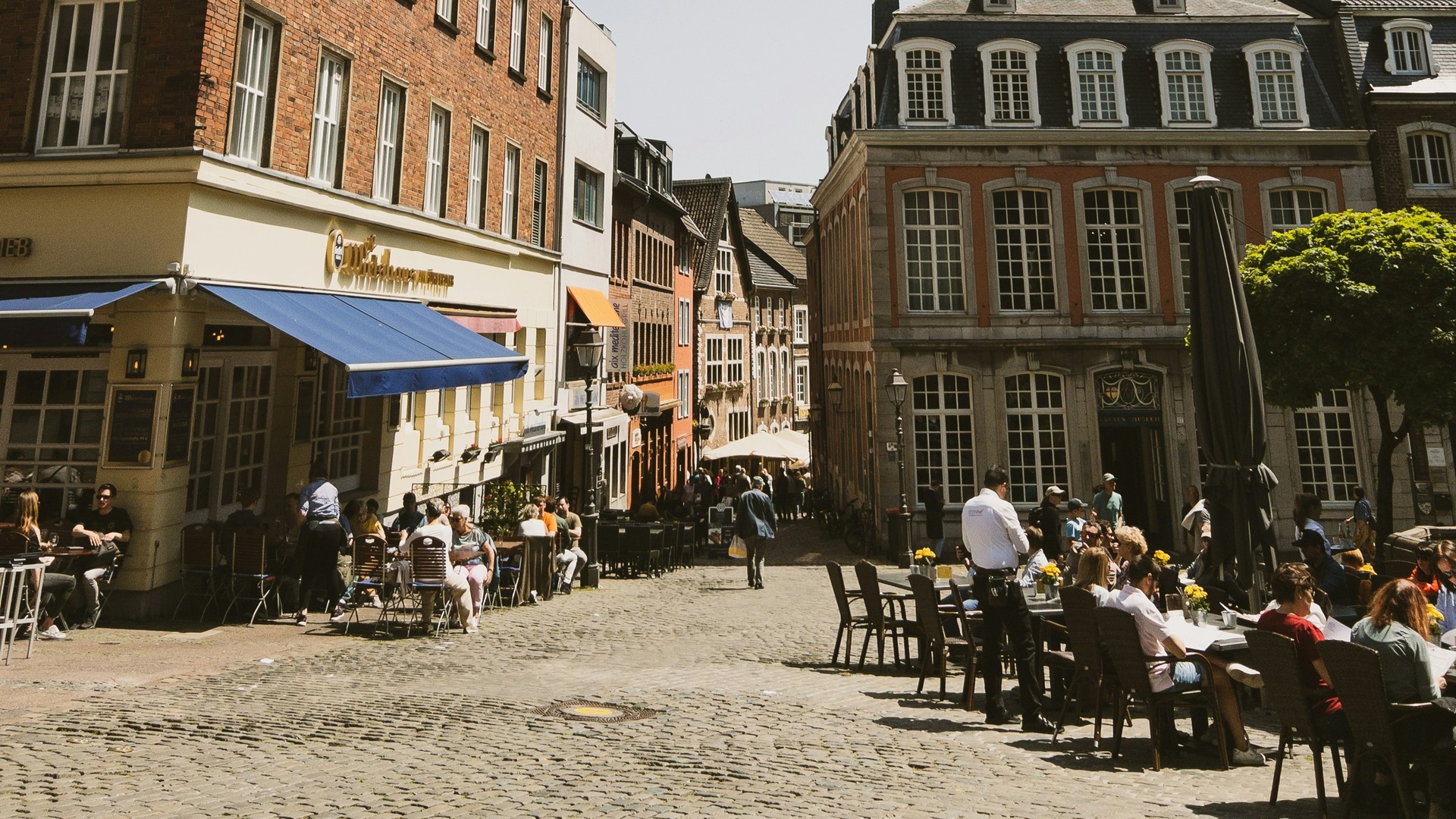 Calle comercial de Colonia con ciclistas, coches aparcados y un rascacielos moderno al fondo.