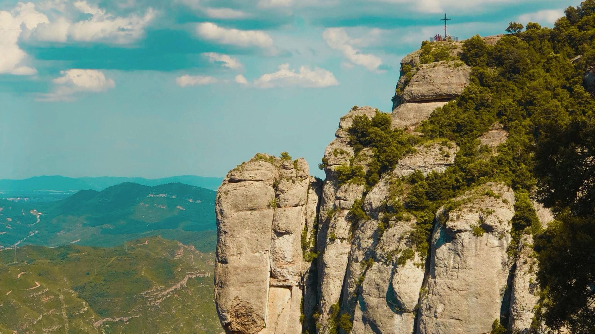 Peñasco de Montserrat con cruz en la cima y montañas verdes al fondo.
