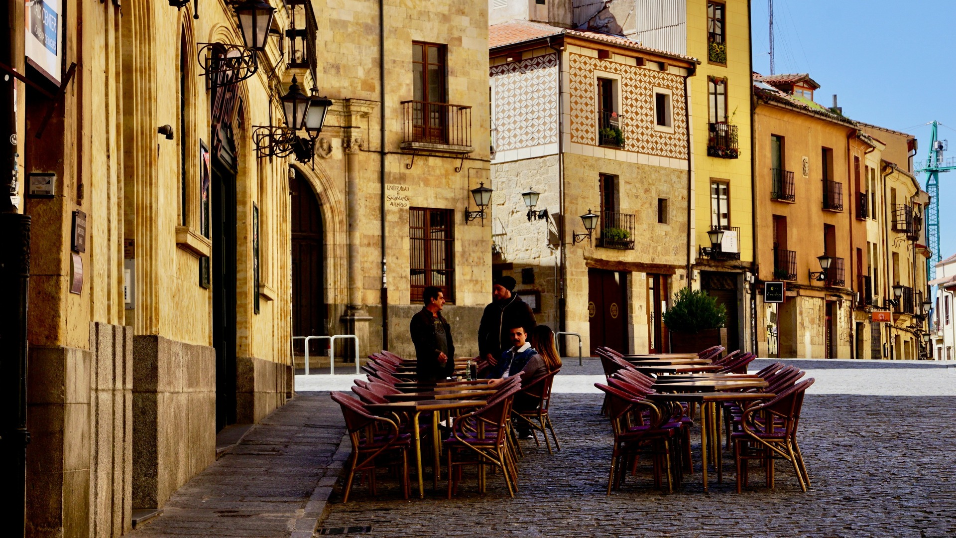 Terraza de café con mesas y sillas en una calle adoquinada del casco antiguo de Salamanca, edificios históricos y sol.