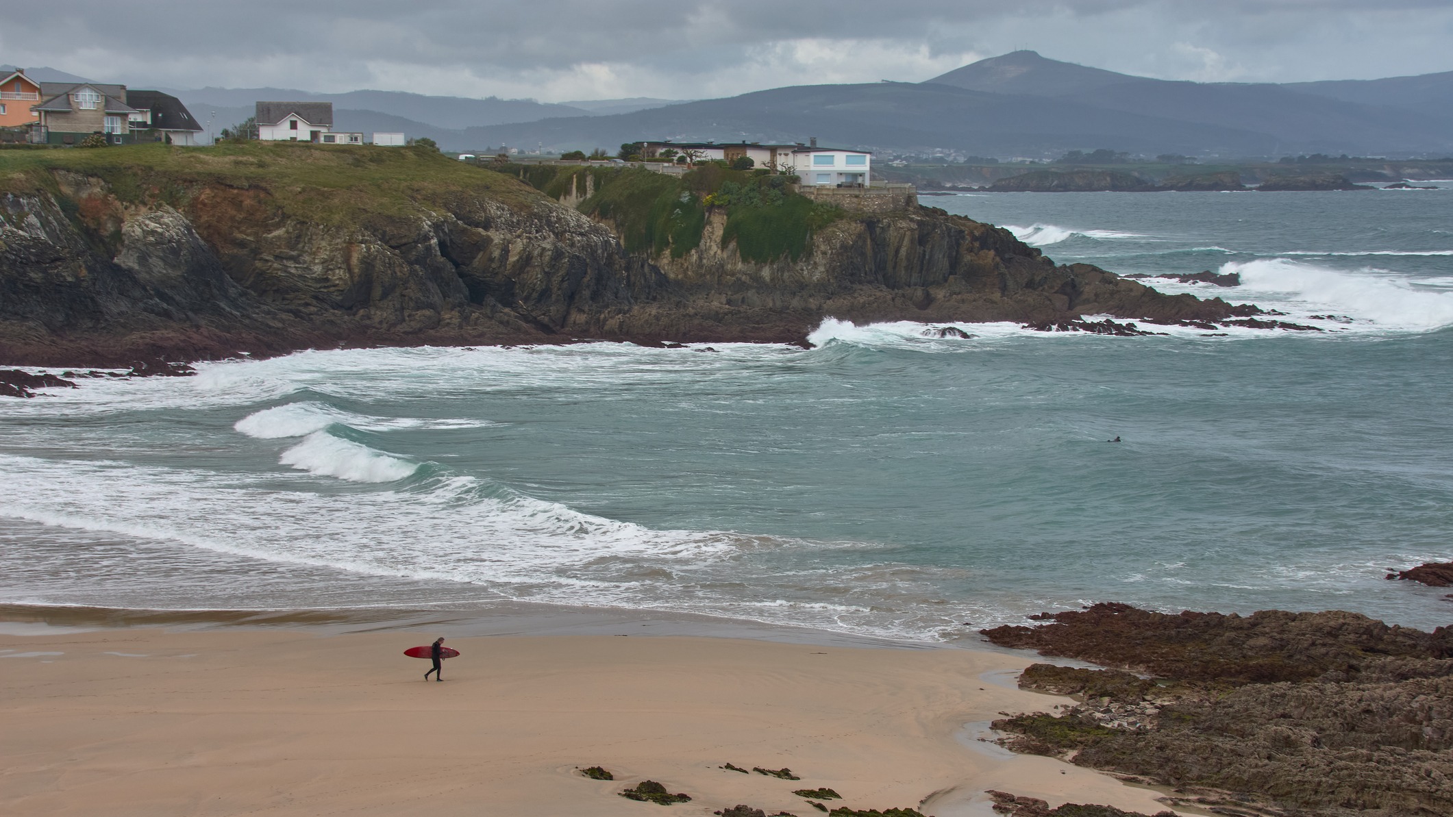 Surfista con tabla roja caminando por la playa de Tapia de Casariego, con el mar Cantábrico y acantilados al fondo.