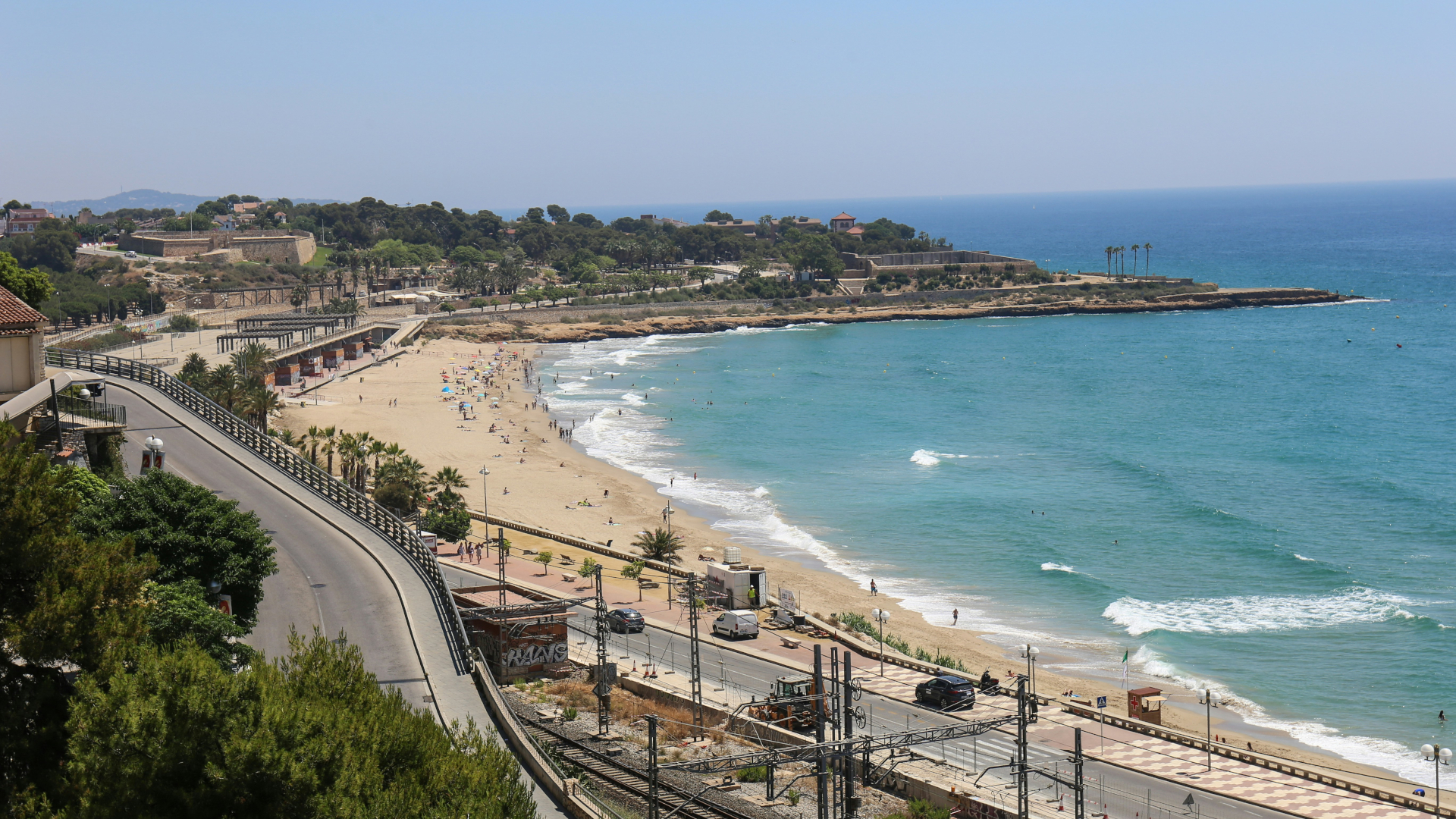 Vista aérea de la Playa del Miracle en Tarragona, con la línea costera, el mar azul y la carretera y tren en primer plano.