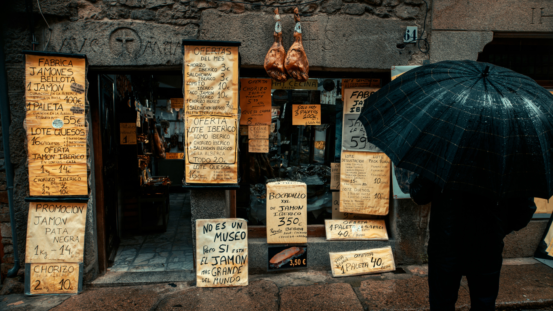 Tienda de jamón ibérico en España con ofertas escritas a mano y jamones colgando, persona con paraguas negro.