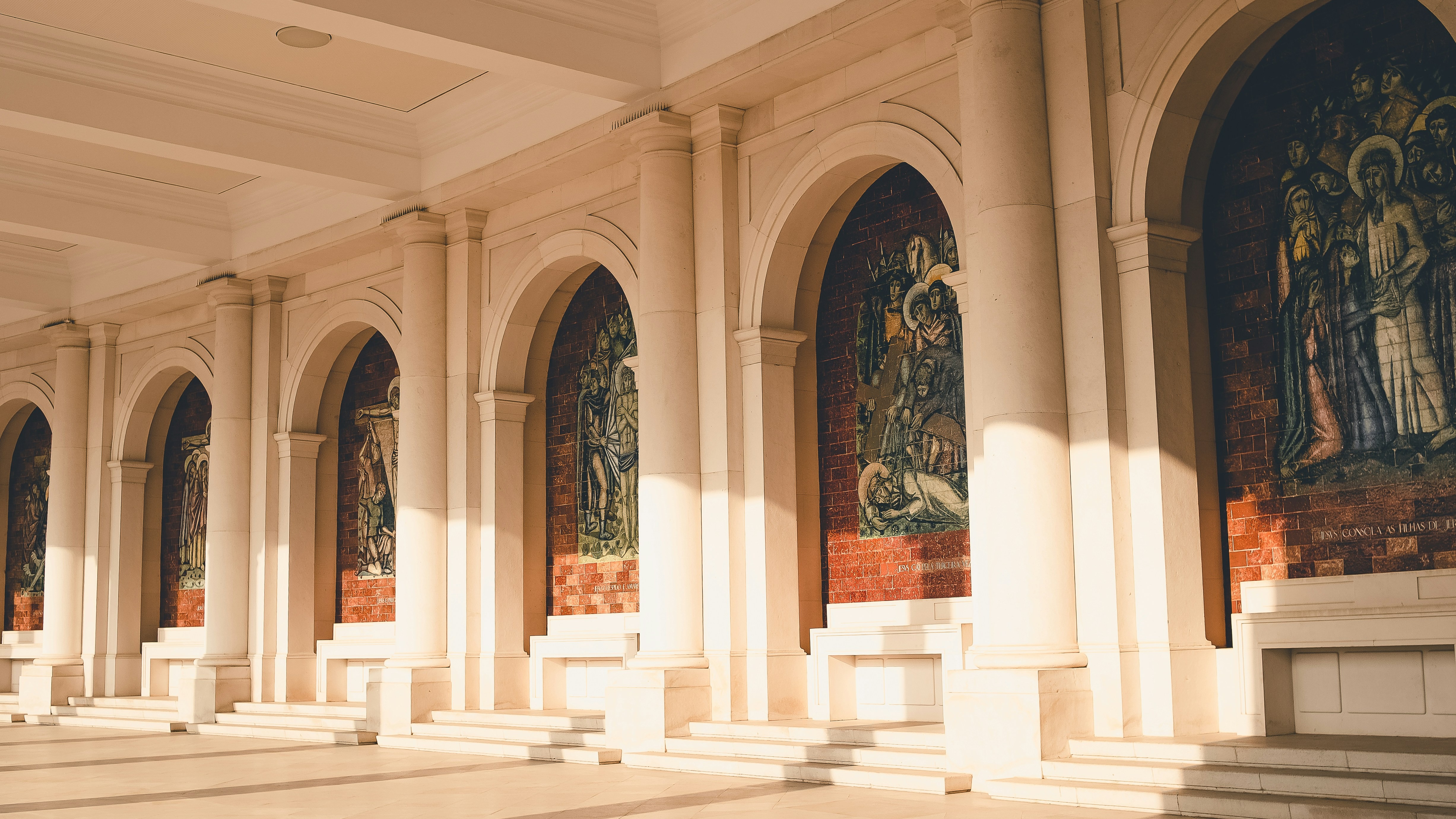 Galerías con arcos y mosaicos religiosos en la columnata del Santuario de Fátima, Portugal, bajo luz dorada.