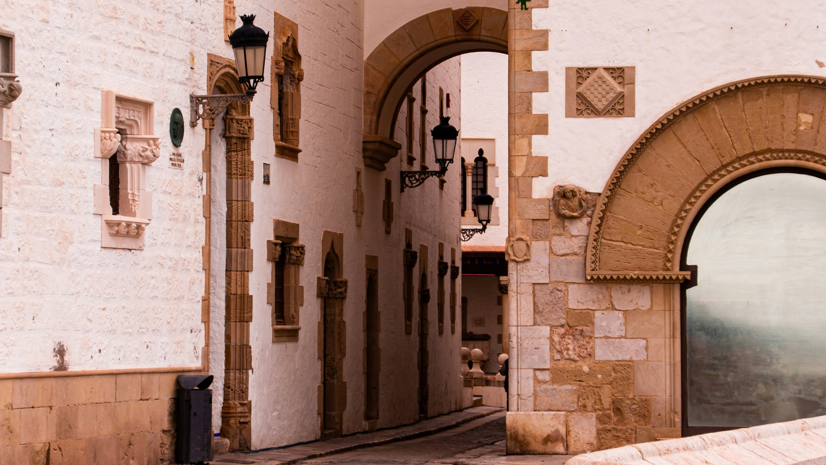 Calle estrecha de Sitges con muros blancos, arcos de piedra y faroles antiguos.