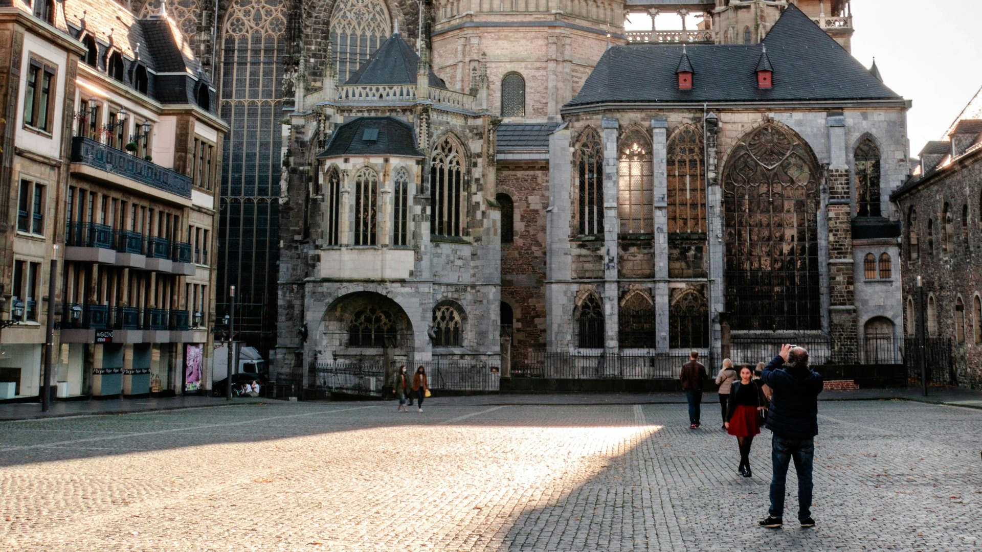  Vista lateral de la Catedral de Aquisgrán con un ábside gótico, en una plaza con gente al sol.