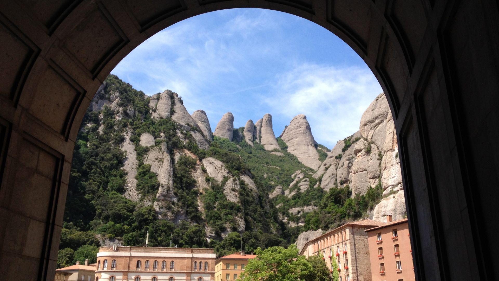 Vista enmarcada por un arco desde el monasterio de Montserrat hacia los picos rocosos, cubiertos de vegetación.