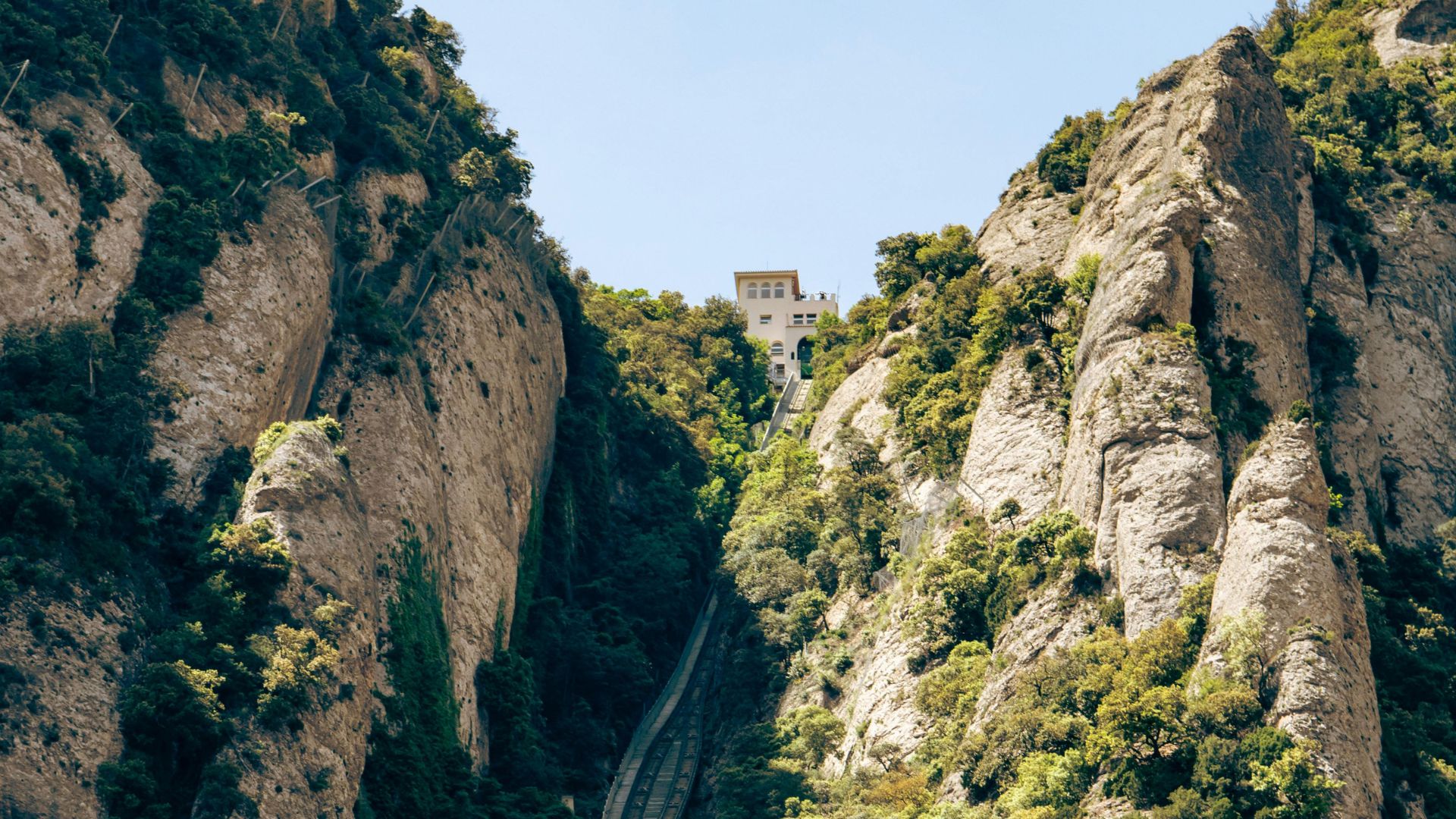 Funicular de Sant Joan subiendo por la grieta de roca de Montserrat, rodeado de vegetación y edificio en la cima.