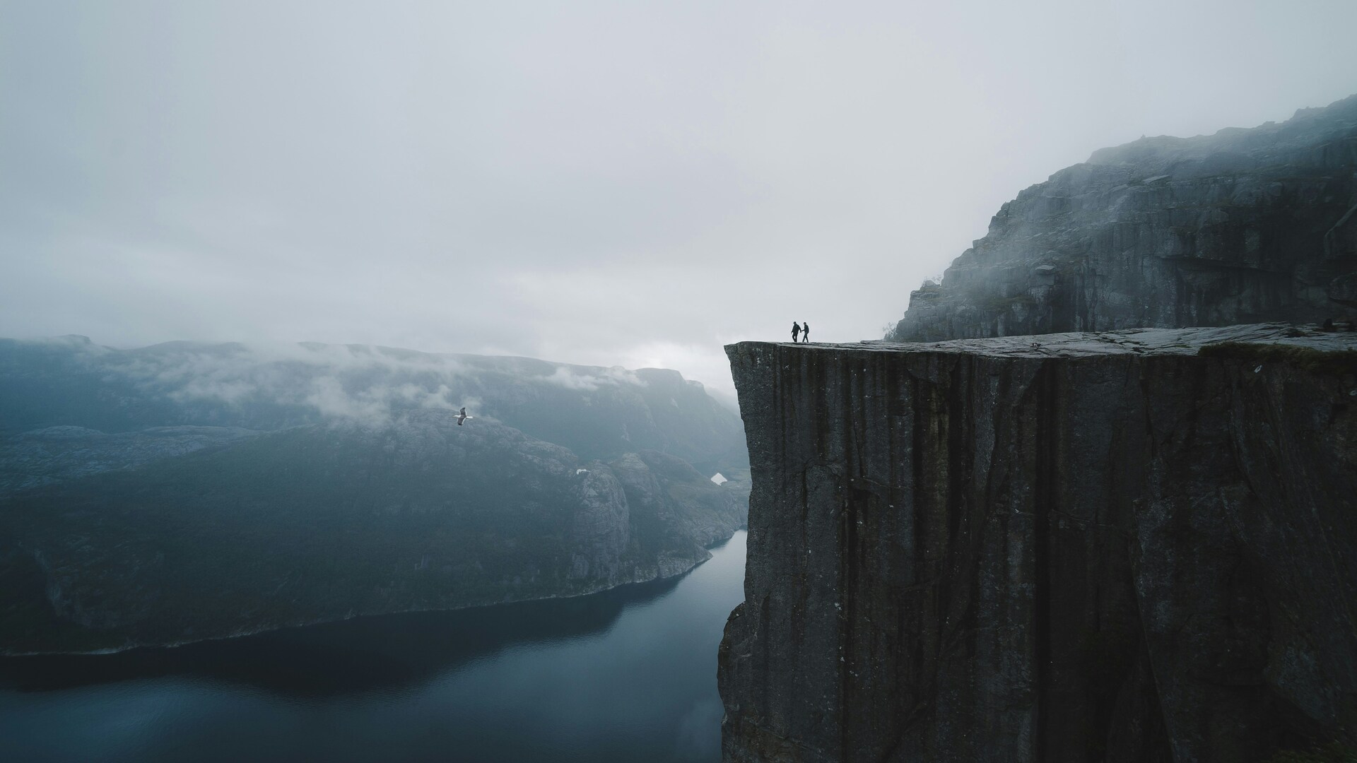 Dos personas en la cima de Preikestolen (Pulpit Rock) sobre el fiordo Lysefjord, Noruega, con niebla y nubes.