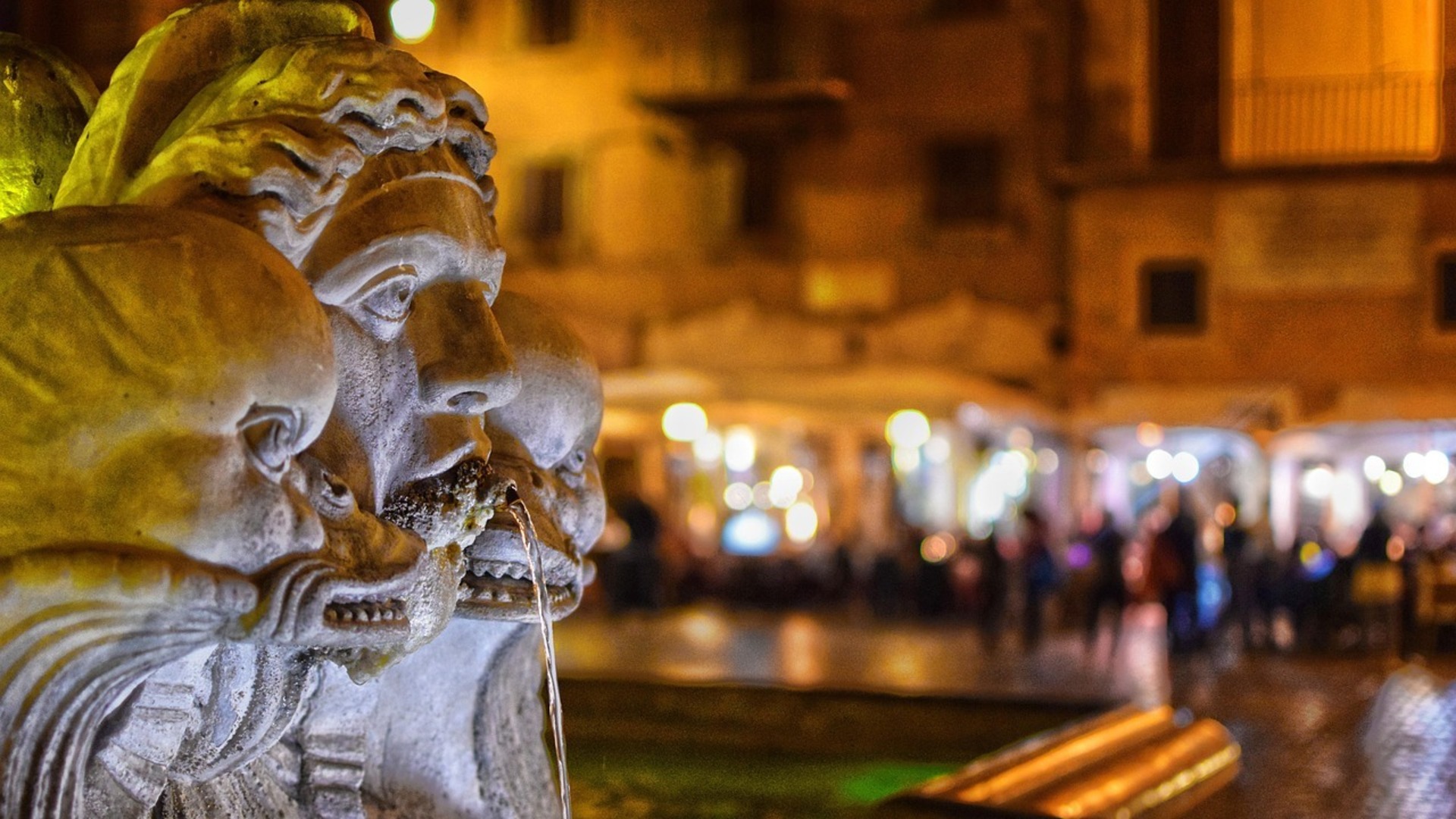 Primer plano de escultura en fuente romana con plaza iluminada al fondo de noche.