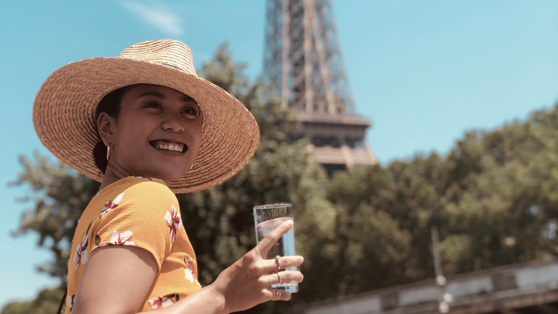 Mujer sonriente con sombrero y bebida frente a la Torre Eiffel en un día soleado.