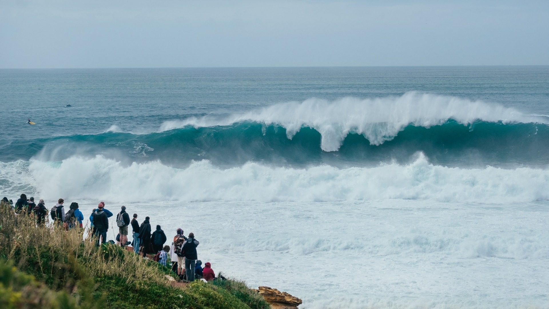 Turistas observando una ola gigante rompiendo en la costa de Nazaré, Portugal.