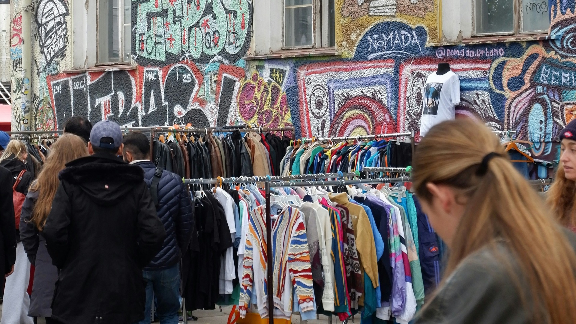 Gente revisando percheros de ropa con grafiti al fondo en Berlín.