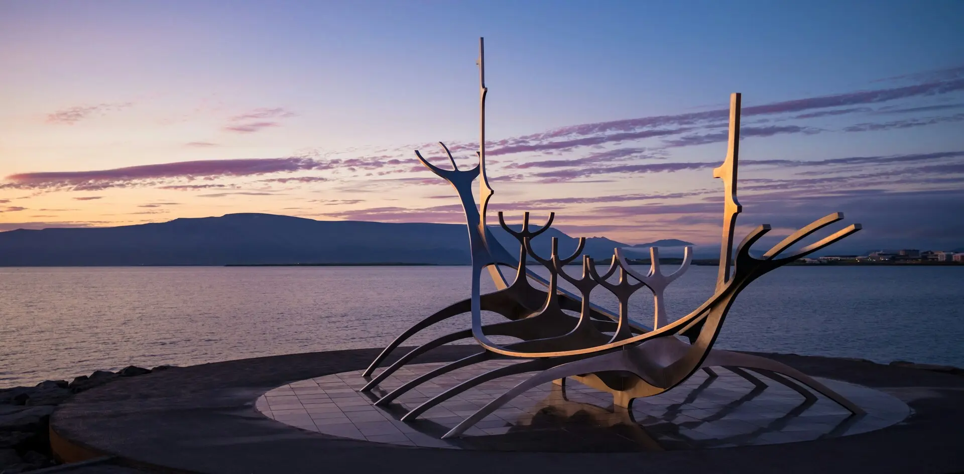 Escultura del Viajero del Sol en Reikiavik al atardecer, con vista al mar y montañas de Islandia iluminadas por el sol. 