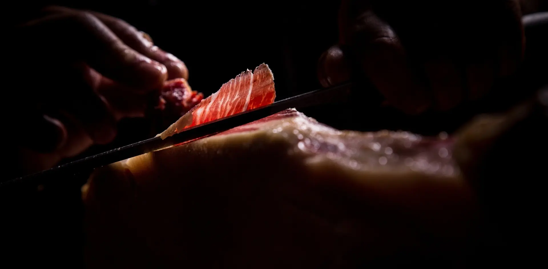 Corte de jamón ibérico con cuchillo, loncha brillante con infiltraciones de grasa, fondo oscuro.