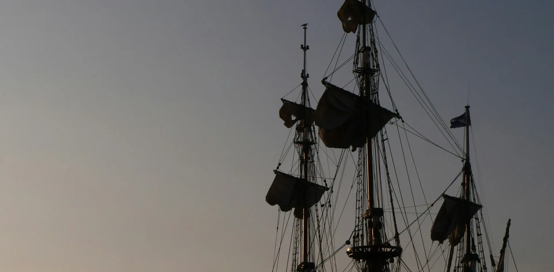 Mástiles y velas de un barco antiguo o réplica al atardecer, siluetas oscuras contra un cielo despejado