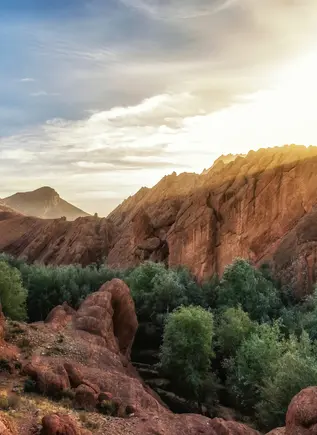 Paisaje del Alto Atlas al atardecer con formaciones rocosas rojizas y un oasis de vegetación frondosa entre las montañas.