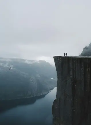 Dos personas en la cima de Preikestolen (Pulpit Rock) sobre el fiordo Lysefjord, Noruega, con niebla y nubes.