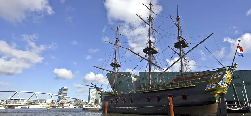 Réplica de un histórico barco de vela neerlandés atracado frente al Museo Marítimo de Ámsterdam, con cielo azul y puentes modernos en el fondo.