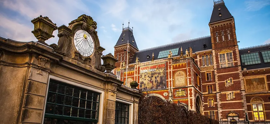 Fachada del Rijksmuseum en Ámsterdam al atardecer, con su arquitectura neorrenacentista de ladrillo rojo y una estructura de reloj solar ornamental en primer plano.