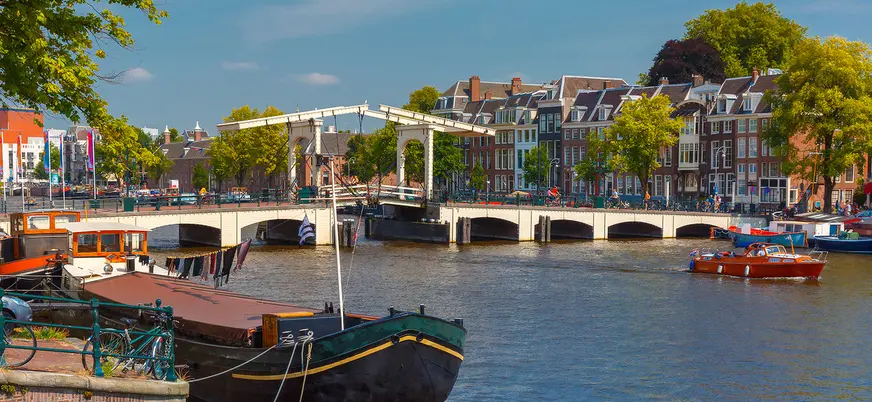 Vista de un canal en Ámsterdam con barcos amarrados y gente navegando, al fondo el puente levadizo blanco Magere Brug y casas típicas holandesas rodeadas de árboles.
