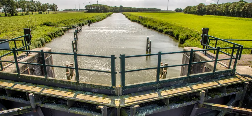 Compuerta y canal de agua en medio de campos verdes, con árboles y turbinas eólicas al fondo bajo un cielo nublado en un paisaje rural neerlandés.