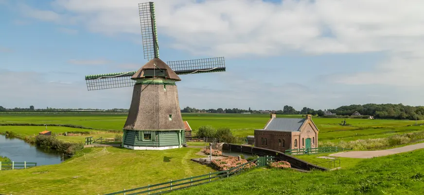 Molino de viento tradicional neerlandés junto a una pequeña casa de ladrillo, rodeados de campos verdes y canales.