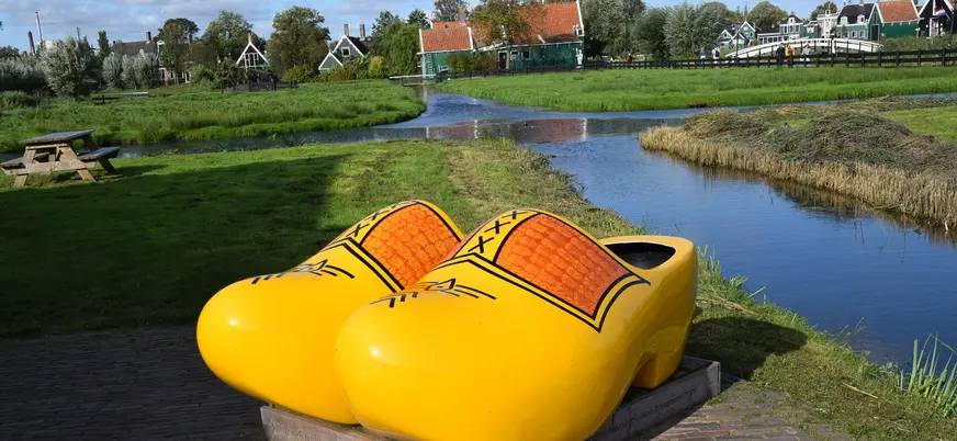 Zuecos de madera gigantes amarillos junto a un canal, con casas tradicionales neerlandesas y molinos de fondo en un paisaje típico de Ámsterdam.