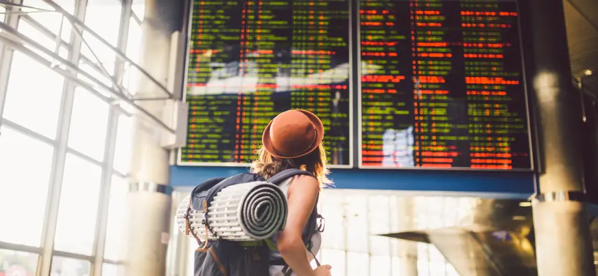 Chica con mochila y sombrero mirando el panel de información de vuelos en un aeropuerto, con listas de salidas y llegadas iluminadas en pantalla.