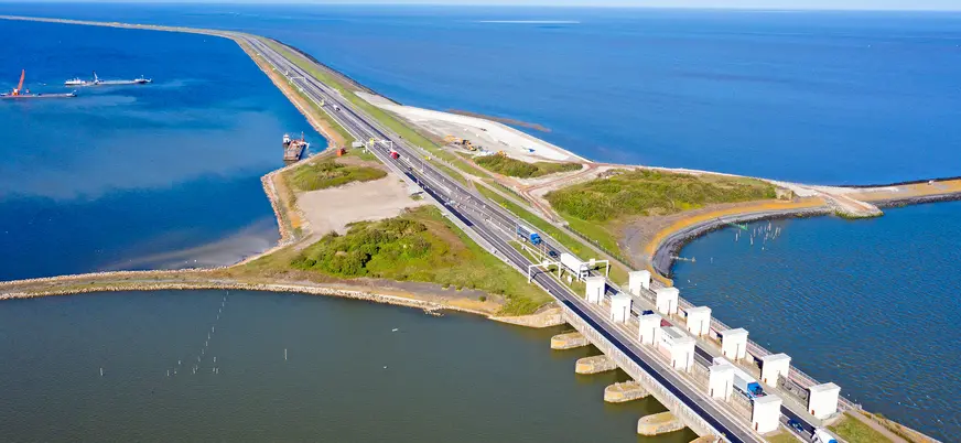 Vista aérea de una carretera y un dique (Afsluitdijk) que separa dos extensas masas de agua en los Países Bajos, con estructuras de control de agua y vehículos circulando, mostrando la ingeniería hidráulica holandesa.