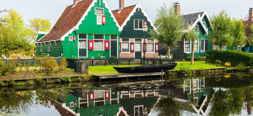 Casas típicas holandesas de madera verde con tejados a dos aguas, situadas junto a un canal en el que se reflejan, rodeadas de árboles y vegetación en Zaanse Schans, Países Bajos.