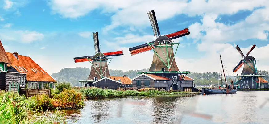 Molinos de viento tradicionales junto a un canal, rodeados de casas con techos rojos y vegetación, bajo un cielo azul con nubes en Zaanse Schans, Países Bajos.