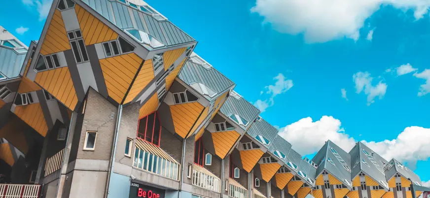 Fila de casas cúbicas amarillas y grises con techos inclinados en Rotterdam, conocidas por su diseño arquitectónico innovador, bajo un cielo azul brillante con algunas nubes blancas.