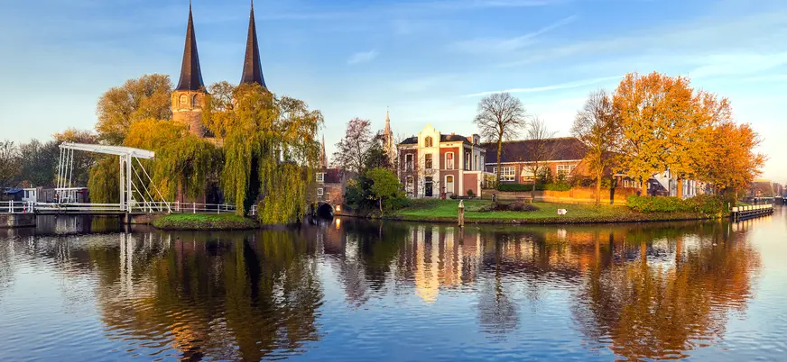 Vista de la Puerta del Este (Oostpoort) en Delft, Países Bajos, con dos altas torres góticas, árboles otoñales y edificios históricos reflejados en el agua tranquila de un canal.