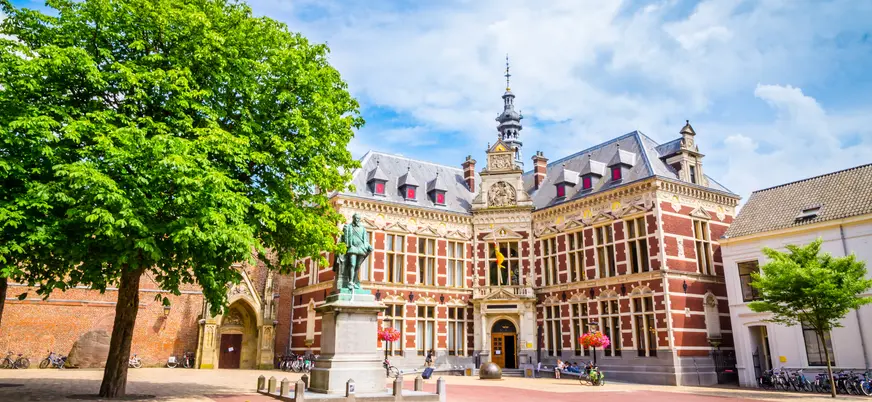 Edificio histórico de la Universidad de Utrecht en los Países Bajos, situado en una plaza abierta con árboles y una estatua en el centro. 