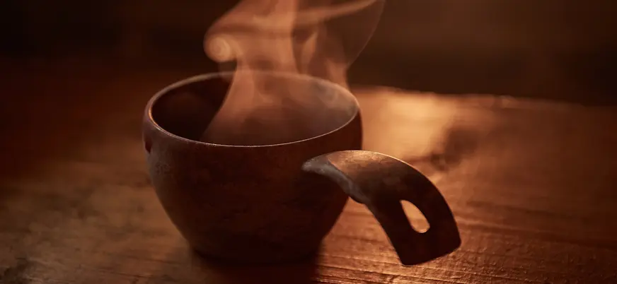 Taza rústica de madera con bebida caliente humeante sobre una mesa de madera iluminada por luz cálida.