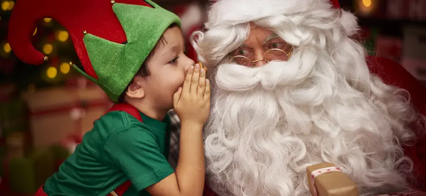 Niño vestido de duende navideño susurrando al oído de Papá Noel, rodeados de regalos en un ambiente festivo típico de Navidad.