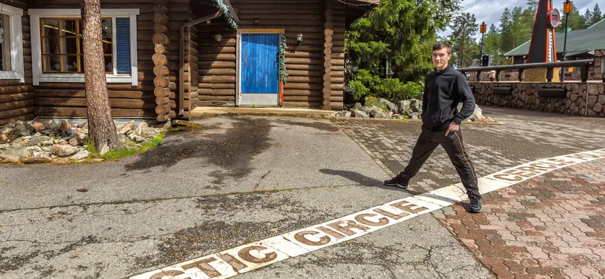 Línea del Círculo Ártico frente a una cabaña de madera con puerta azul en un entorno natural, rodeado de árboles y rocas.